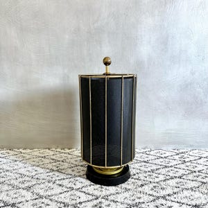 Könnte beinhalten: Eine Vintage-Lampe in Schwarz und Gold mit einem zylindrischen Schirm. Der Schirm hat ein perforiertes Metalldesign und wird von vertikalen Goldstäben umrahmt. Die Lampe steht auf einem schwarzen Sockel mit goldenen Akzenten und einer kleinen, runden Spitze.