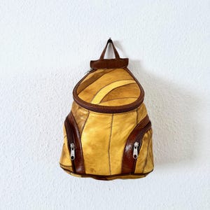 Könnte beinhalten: Vintage Rucksack aus gelbem und braunem Leder. Die Tasche hat einen abgerundeten Deckel mit Reißverschluss, zwei Vordertaschen mit Reißverschlüssen und einen kleinen Griff. Das Leder ist in einem Patchwork-Design zusammengenäht. Die Tasche hängt an einer weißen Wand.