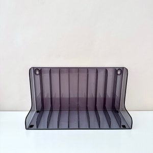 May include: A clear, grey plastic storage rack with 10 compartments for organising items. The rack is designed to hold small items like game cartridges or other collectibles.
