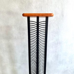 May include: A tall, black metal stand with a rectangular, light brown wooden top. The stand features a repeating, curved design. The stand is approximately 30 inches tall.