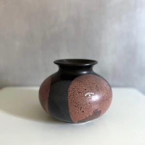 Könnte beinhalten: Eine runde Keramikvase mit dunkelbraunem Rand und gesprenkeltem Körper. Der Körper ist in drei vertikale Abschnitte aus Schwarz, Grau und Rotbraun unterteilt. Auf der Seite ist ein kleiner weißer Aufkleber zu sehen.