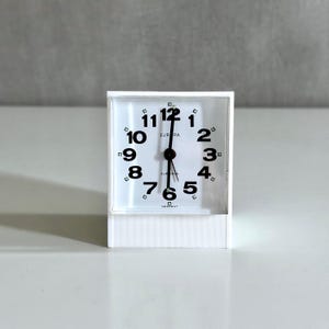 Könnte beinhalten: Ein weißer quadratischer Europa-Wecker mit schwarzen Ziffern und Zeigern. Das Zifferblatt ist weiß mit dem schwarzen Aufdruck "EUROPA". Die Uhr wird in Deutschland hergestellt.