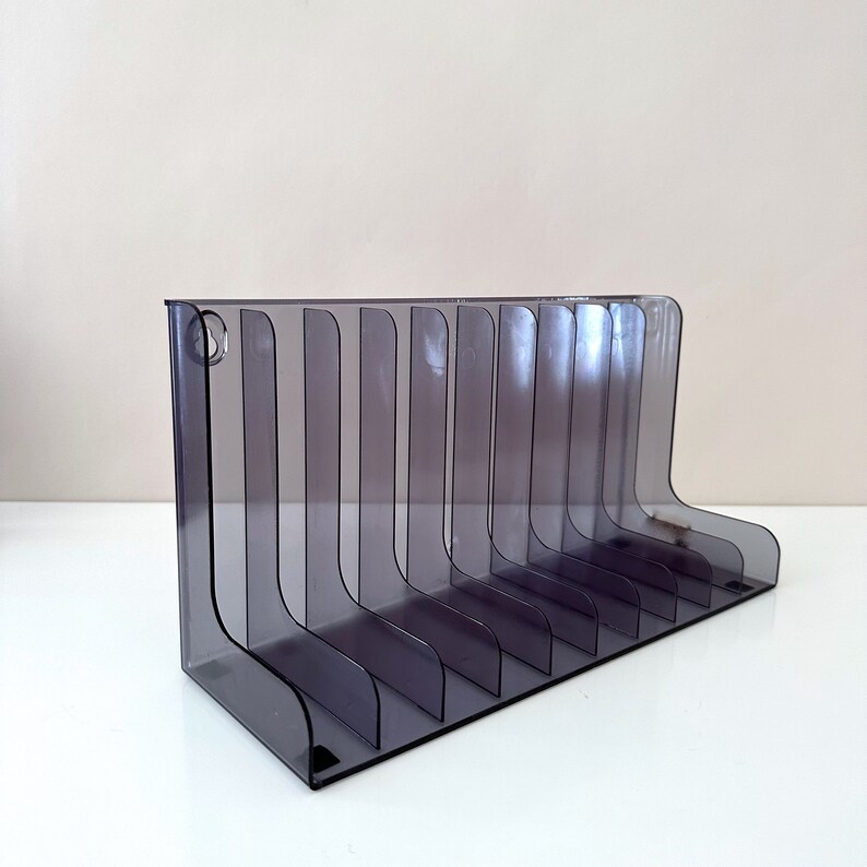 May include: A clear, grey plastic magazine rack with 10 slots for holding magazines or other items. The rack is designed to stand upright on a flat surface.