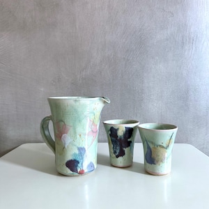 Könnte beinhalten: Ein Keramikkrug und zwei passende Tassen. Der Krug hat einen Henkel und einen Ausguss, und alle drei Teile sind mit einem Aquarell-Blumenmuster in Rosa-, Blau- und Lilatönen auf hellgrünem Hintergrund bemalt.