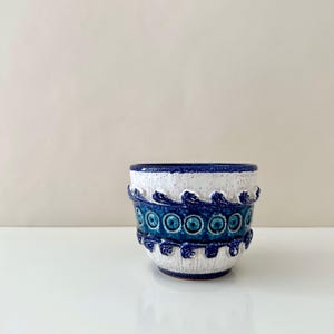 May include: A ceramic planter with a white speckled top and a band of blue and white wave-like designs. The middle section features a series of blue circles with a smaller circle in the center. The planter is a decorative item.