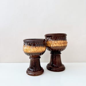 Puede incluir: Dos maceteros de pedestal de cerámica marrón. Los maceteros tienen una base y un borde marrón oscuro, con un diseño floral marrón claro alrededor del centro. Los maceteros son de diferentes tamaños.