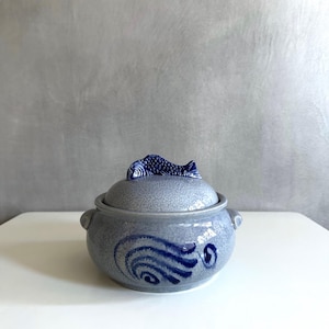 May include: A blue and white ceramic pot with a fish-shaped lid. The pot has a blue and white wave design.