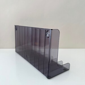 May include: A clear, grey plastic wall-mounted organiser with multiple slots for storing items. The organiser has a slight curve at the bottom and is designed to be mounted on a wall.