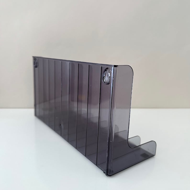 May include: A clear, grey plastic wall-mounted organiser with multiple slots for storing items. The organiser has a slight curve at the bottom and is designed to be mounted on a wall.