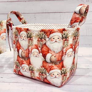 May include: A fabric storage basket with a festive Santa Claus print. The basket features a repeating pattern of Santa Claus figures in red suits, white beards, and red hats. The handles and interior lining have a complementary design. The basket is designed for storage.