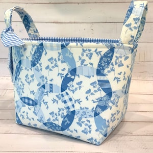 May include: A fabric storage basket with a blue and white floral pattern. The basket has two handles and a gingham trim. The exterior features a patchwork design with floral and geometric elements. The interior is lined with a blue and white gingham fabric.