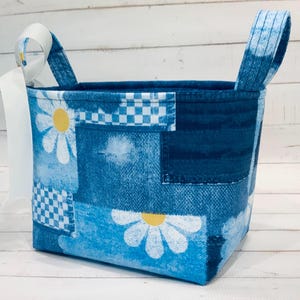 May include: A fabric storage basket featuring a patchwork design of blue denim-style textures, white daisy flowers with yellow centers, and blue and white checkered patterns. The basket has two upright fabric handles and a white ribbon tied to one side.