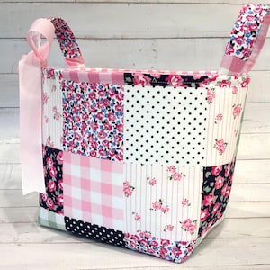 May include: A fabric storage basket featuring a patchwork design of pink and white gingham, black and white polka dots, and floral patterns with pink roses on dark blue and white backgrounds. It includes two fabric handles and a light pink ribbon.