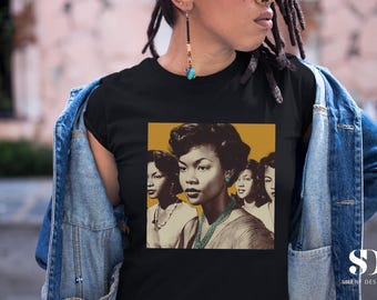 May include: Black t-shirt with a vintage-style portrait of several women with dark hair and skin. The portrait is set against a mustard-yellow background. The shirt is worn with a denim jacket.
