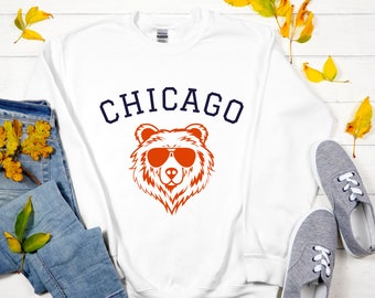 Może przedstawiać: Biała bluza z nadrukiem "CHICAGO" w kolorze granatowym i pomarańczowym niedźwiedziem w okularach przeciwsłonecznych.