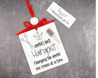 May include: A white metal hairdresser ornament with a red ribbon and a white bead. The ornament has a drawing of scissors, a crown, and a hairbrush. Text on the ornament reads "world's best Hairapist Changing the world one crown at a time."