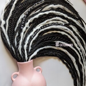 May include: Black and white dreadlocks styled in a vase-like pink ceramic container. The dreadlocks are a mix of black and white strands, with white stitching details. The text "BB EXTENSIONS" is visible.