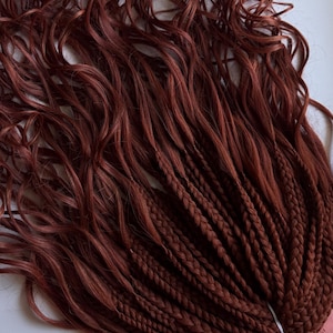 May include: A bundle of auburn-colored synthetic hair extensions. The hair is a mix of wavy and braided styles, with a transition from wavy at the top to neatly braided at the bottom. The extensions are tied together with a white band.