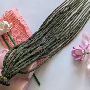 May include: A set of dark green synthetic dreadlocks, with a pink and white flower on the left and a pink flower on the right.