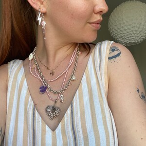 May include: A woman wearing a striped tank top with multiple necklaces, including a heart-shaped pendant, a pearl necklace, and a chain with a charm. She is also wearing earrings with a lightning bolt charm and a small floral charm.