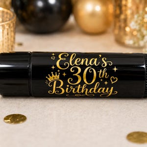 May include: A black cylindrical container with gold text that reads "Elena's 30th Birthday". The text is embellished with stars, a crown, and hearts. The background includes gold confetti and decorative items.