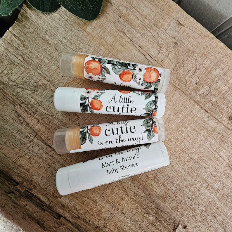 May include: Four white tubes of lip balm with orange and floral designs. The labels read "A little cutie is on the way!" and "Matt & Anna's Baby Shower".