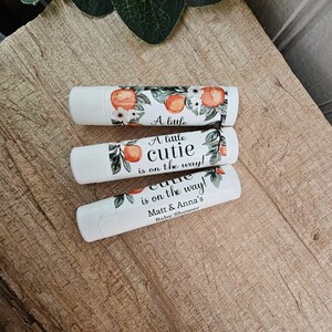 May include: Three white lip balm tubes with orange blossom and leaf designs. The labels read "A little cutie is on the way!" and "Matt & Anna's Baby Shower".