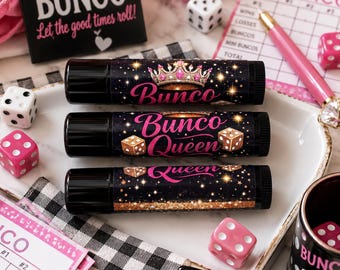 May include: Three black lip balm tubes featuring gold and pink graphics. The top tube displays a crown and the text Bunco. The middle tube features the text Bunco Queen and dice illustrations. The bottom tube shows the text Queen with gold glitter.