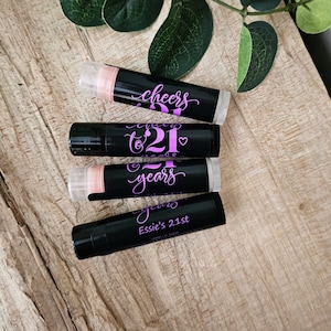 May include: Five black lip balm tubes with pink and white lettering. The tubes are labeled "Cheers to 21 years" and "Essie's 21st".