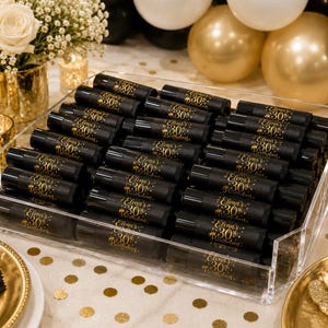 May include: A clear acrylic tray filled with black tubes, each labeled with gold text reading "Elena's 30th Birthday." The tubes are arranged on a white tablecloth with gold confetti. The background includes gold and white balloons, flowers, and candles.