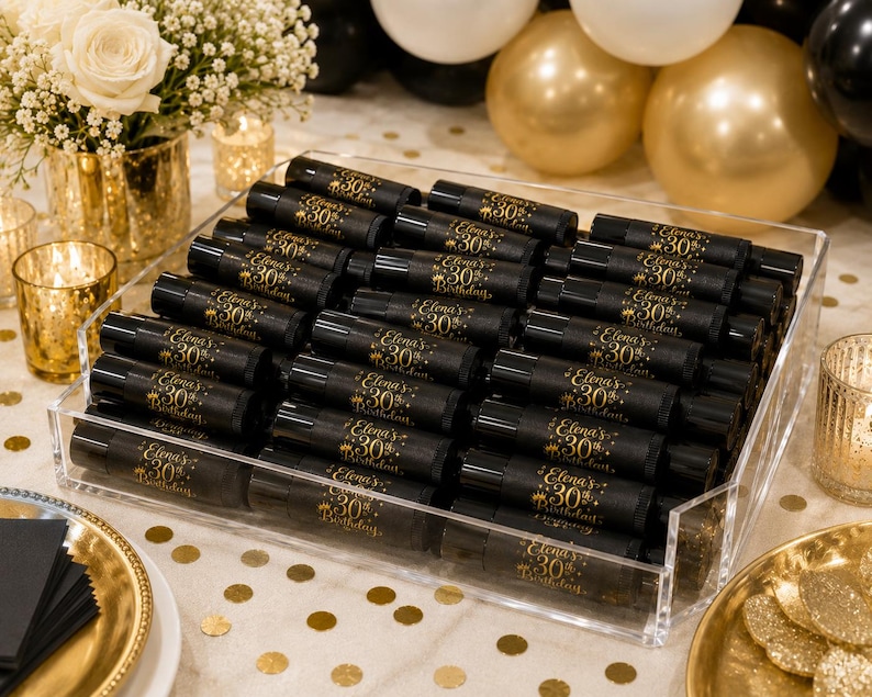 May include: A clear acrylic tray filled with black tubes, each labeled with gold text reading "Elena's 30th Birthday." The tubes are arranged on a white tablecloth with gold confetti. The background includes gold and white balloons, flowers, and candles.