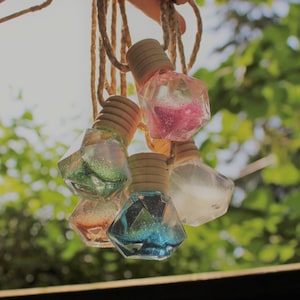 May include: Five decorative glass bottles with glitter and cork tops hanging from twine. The bottles are shaped like diamonds and are filled with different colored glitter.