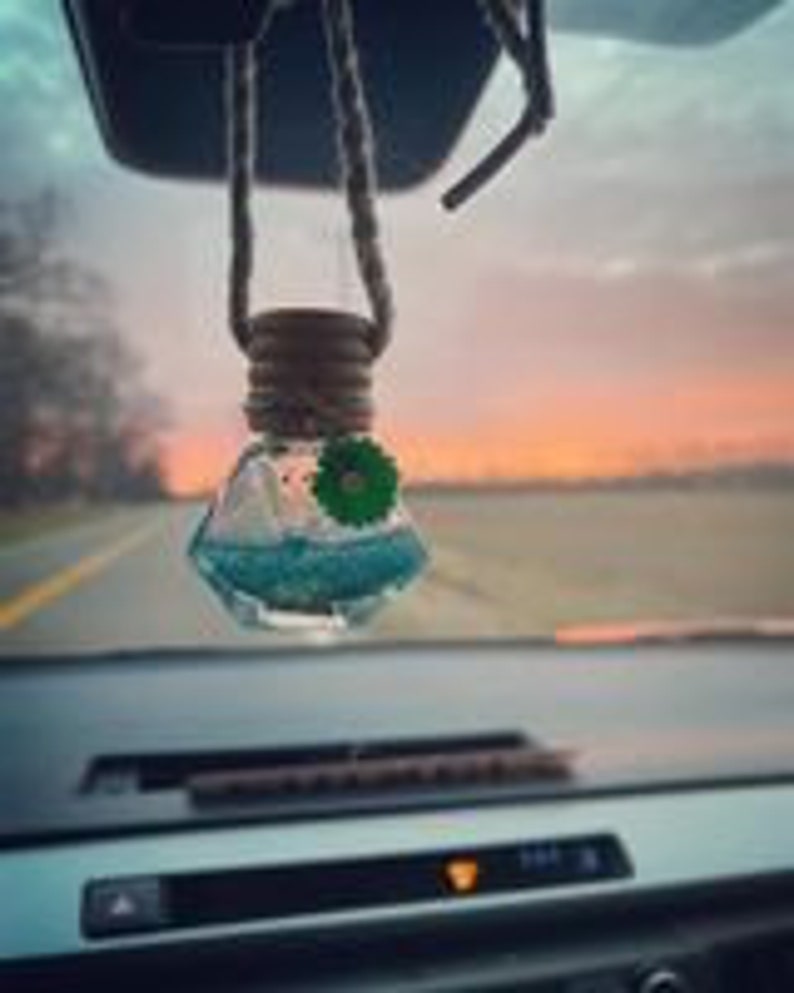 May include: A blue and green car air freshener hanging from a rearview mirror. The air freshener is in a clear, diamond-shaped bottle with a wooden cap and a string.
