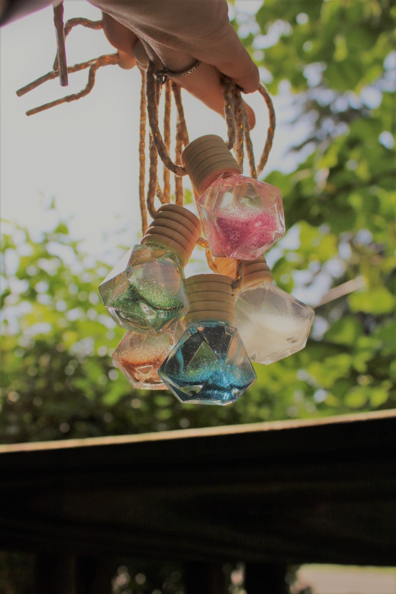 May include: Five decorative glass bottles with glitter and cork tops hanging from twine. The bottles are shaped like diamonds and are filled with different colored glitter.