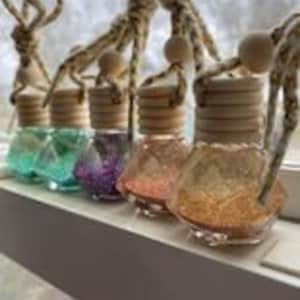 May include: Five small glass bottles with glitter inside and wooden stoppers. The bottles are hanging from a string with a wooden bead. The bottles are in the colors teal, purple, pink, and gold.