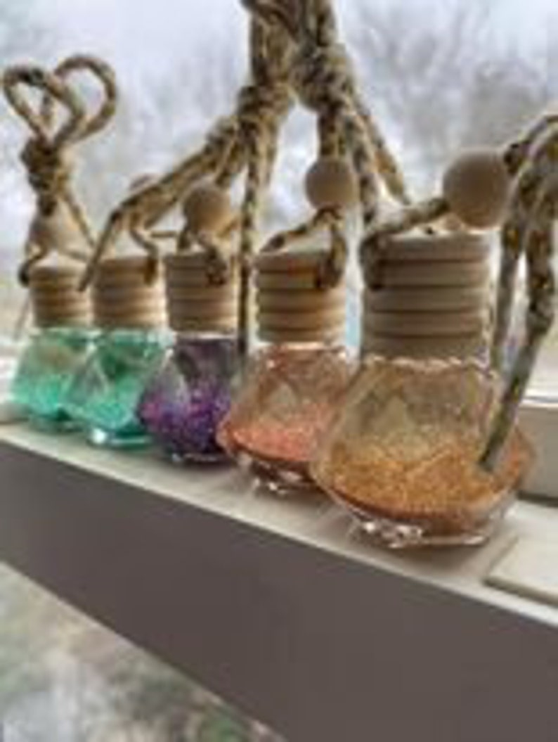 May include: Five small glass bottles with glitter inside and wooden stoppers. The bottles are hanging from a string with a wooden bead. The bottles are in the colors teal, purple, pink, and gold.