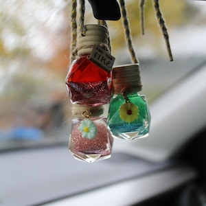 May include: Three glass bottles with glitter and small flower charms hanging from a rearview mirror. The bottles are red, blue, and pink. The red bottle has a tag that says "Find Your Fire".