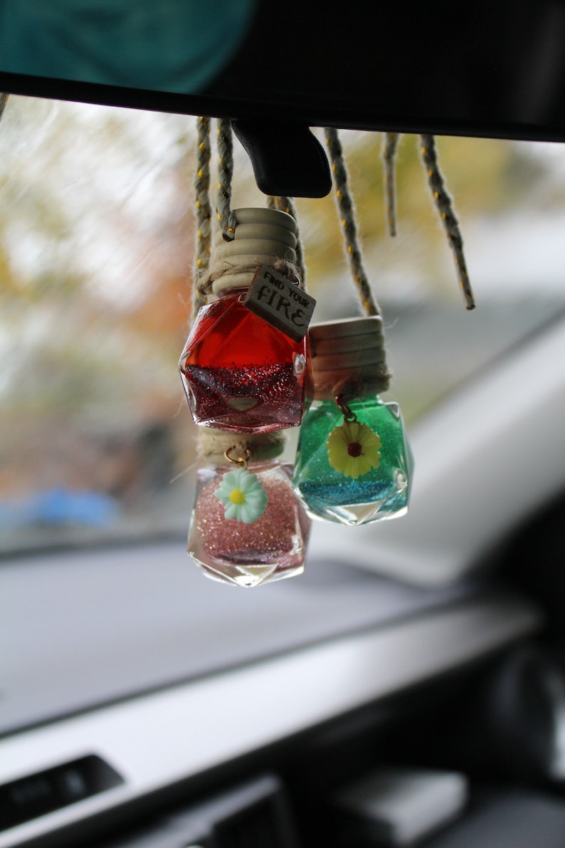 May include: Three glass bottles with glitter and small flower charms hanging from a rearview mirror. The bottles are red, blue, and pink. The red bottle has a tag that says "Find Your Fire".
