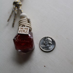 May include: A red glass bottle with a wooden tag that says "Find Your Fire" hanging from a white and gold string. The bottle is shaped like a gemstone and has a glittery finish. A US quarter is included for size reference.
