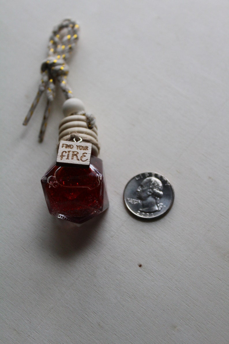 May include: A red glass bottle with a wooden tag that says "Find Your Fire" hanging from a white and gold string. The bottle is shaped like a gemstone and has a glittery finish. A US quarter is included for size reference.