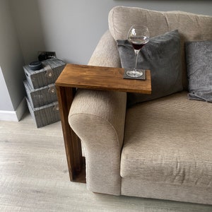 May include: A dark stained wooden sofa arm table. The table is attached to the arm of a beige sofa. A wine glass with red wine sits on the table, along with a coaster. Grey cushions are behind the table.