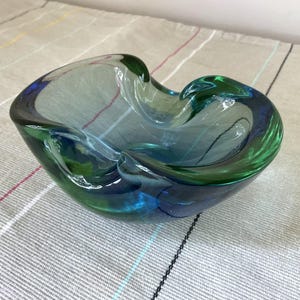 May include: A free-form glass bowl featuring a blend of translucent blue and green hues. The vessel has an organic, asymmetrical shape with thick, smooth walls and a polished finish, showcasing a swirling colour gradient throughout the glass.