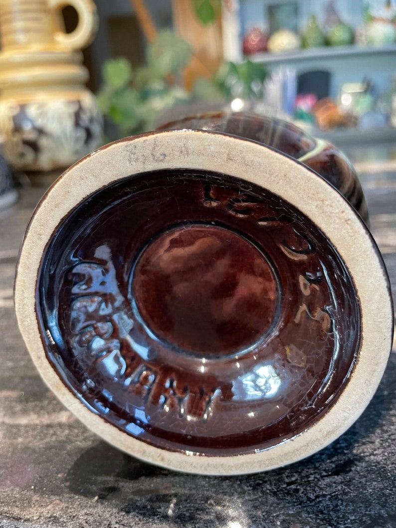 May include: A brown ceramic vase with a white rim and a brown glaze. The bottom of the vase has a white circle with the text "Germany" in black.