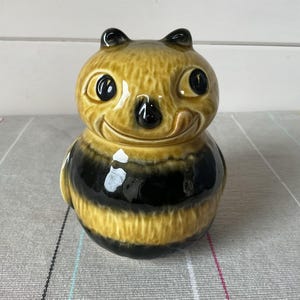 May include: A ceramic bee-shaped jar with a smiling face, black eyes, and antennae. The jar has a yellow and black striped body, and is likely used for storage. The jar is sitting on a grey surface.