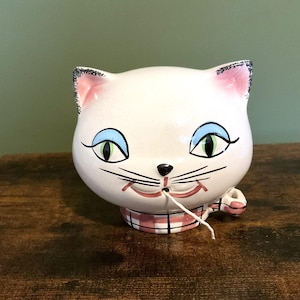 May include: A white ceramic cat-shaped container with pink ears, blue eyes, and a black nose and whiskers. The cat has a pink and white checkered collar. The container is sitting on a wooden surface.