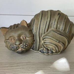 May include: A ceramic cat figurine in a seated position with a muted olive green and brown glaze. The sculpture features dark vertical stripes along the body, textured circular markings on the muzzle, and two almond-shaped eyes.