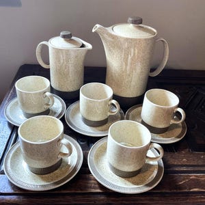 May include: A vintage speckled ceramic coffee set. The set includes a tall coffee pot, a creamer, and six cups with saucers. The cups and saucers have a brown band at the base. The set is beige with brown speckles.