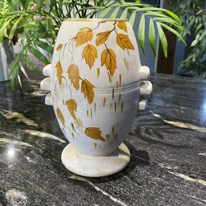 May include: A cream-coloured ceramic vase with a speckled texture, featuring a hand-painted pattern of golden-yellow autumn leaves and vertical brushstrokes. The vase has a rounded body, a pedestal base, and three small horizontal handles on each side.