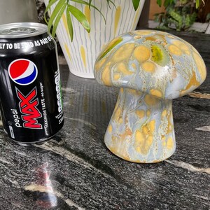 May include: A ceramic mushroom figurine with a yellow and blue glaze. The mushroom has a wide, flat cap and a rounded stem. The figurine is sitting on a black surface.
