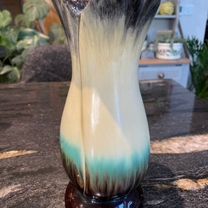 May include: A ceramic vase with a unique design. The vase is white with black and brown accents and a teal blue band around the middle. The vase has a wide mouth and a narrow base.